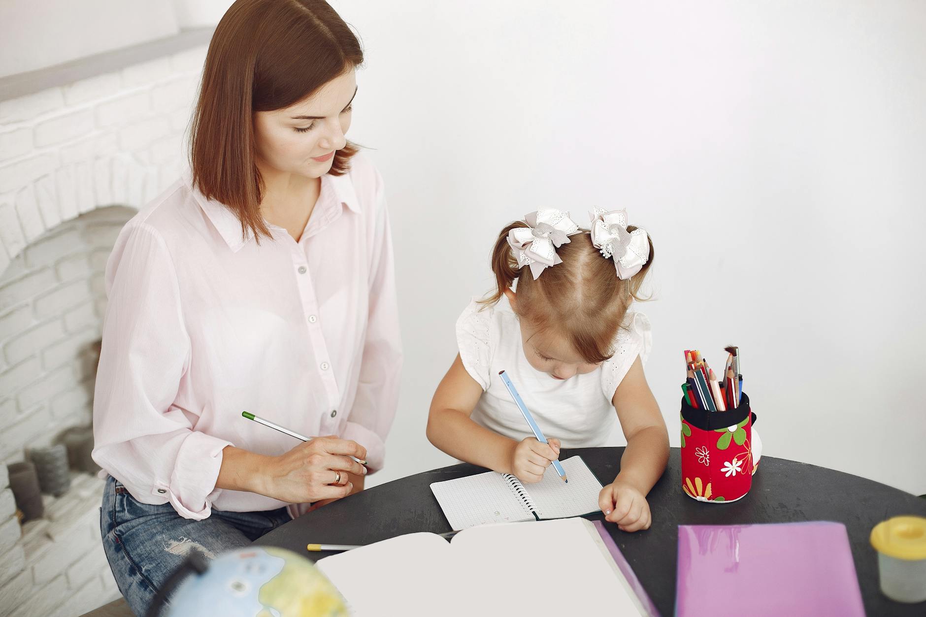 From above small girl drawing with pencil in notepad while mother sitting near at round table
