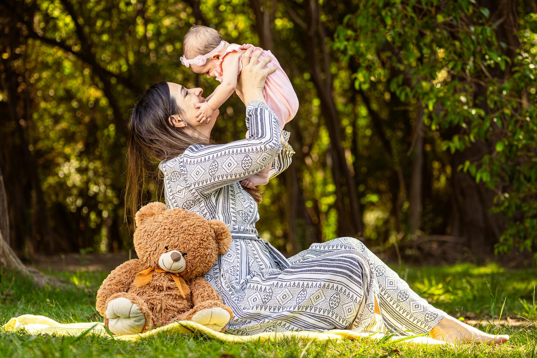 A joyful mother lifting her baby in an outdoor setting with a teddy bear nearby.