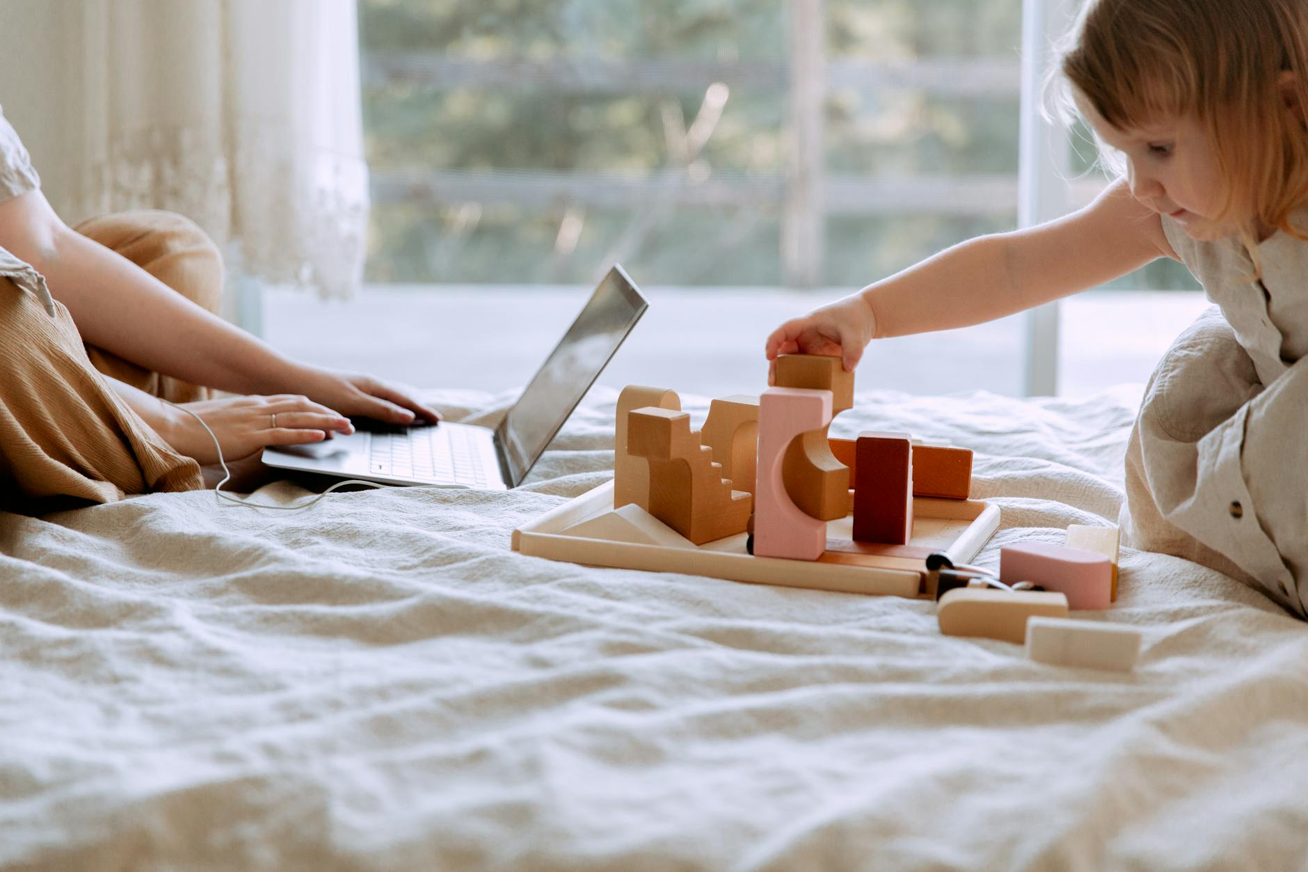Crop woman using laptop while sitting on bed in bedroom with little daughter playing with wooden blocks