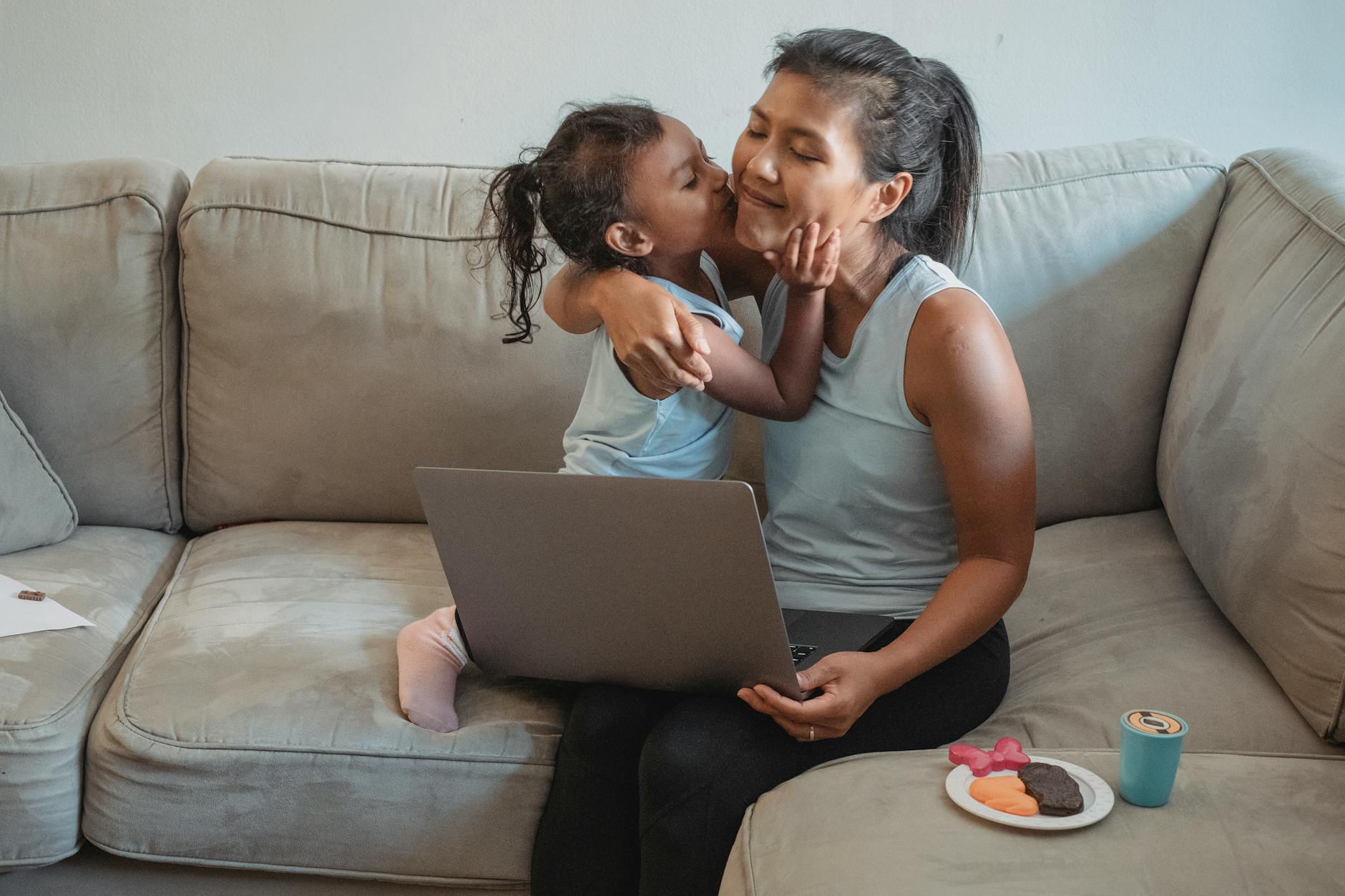 Woman working remotely on netbook while sitting on sofa and cuddling with child near plastic food
