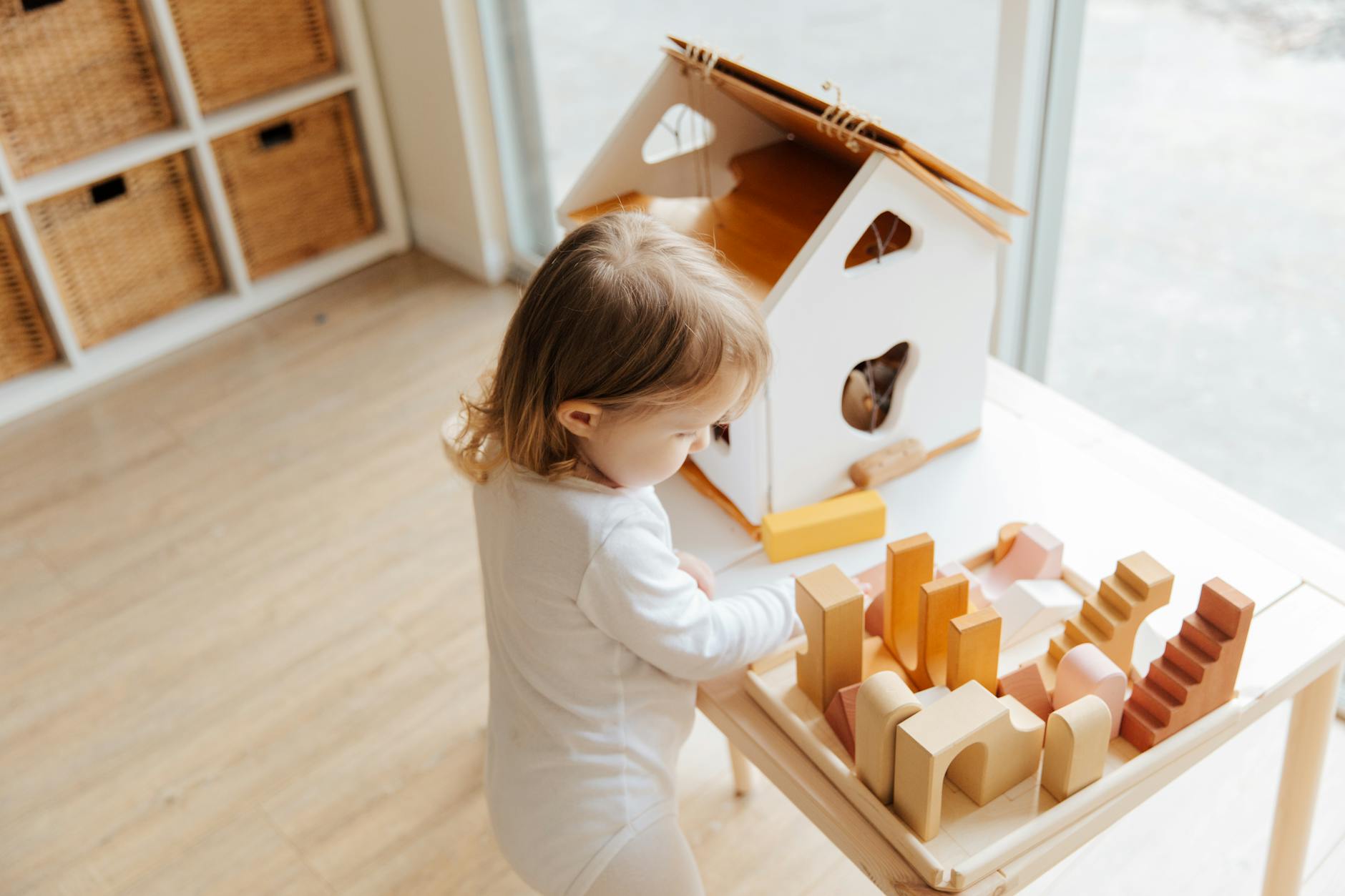 From above view of cute little girl in casual white clothes playing with wooden blocks and toy house at table near window at home