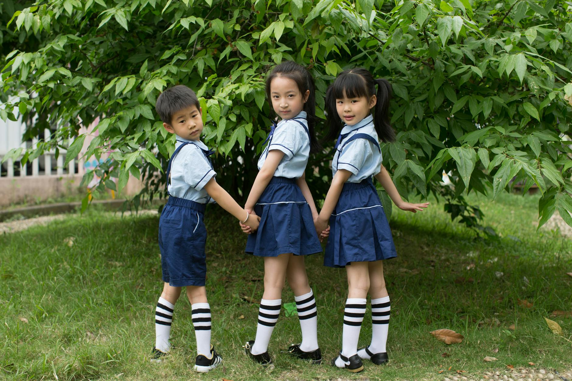 Three children in school uniforms holding hands outdoors, smiling at the camera.