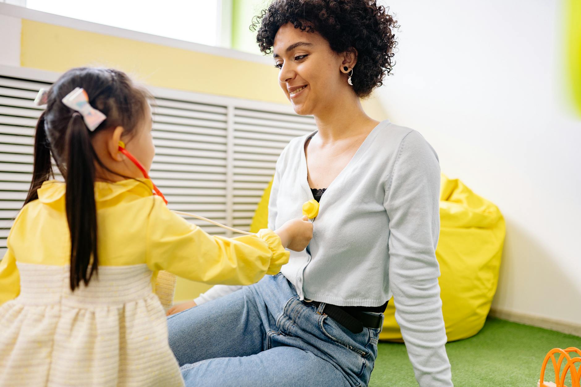 A child plays doctor with a stethoscope and a smiling adult indoors.