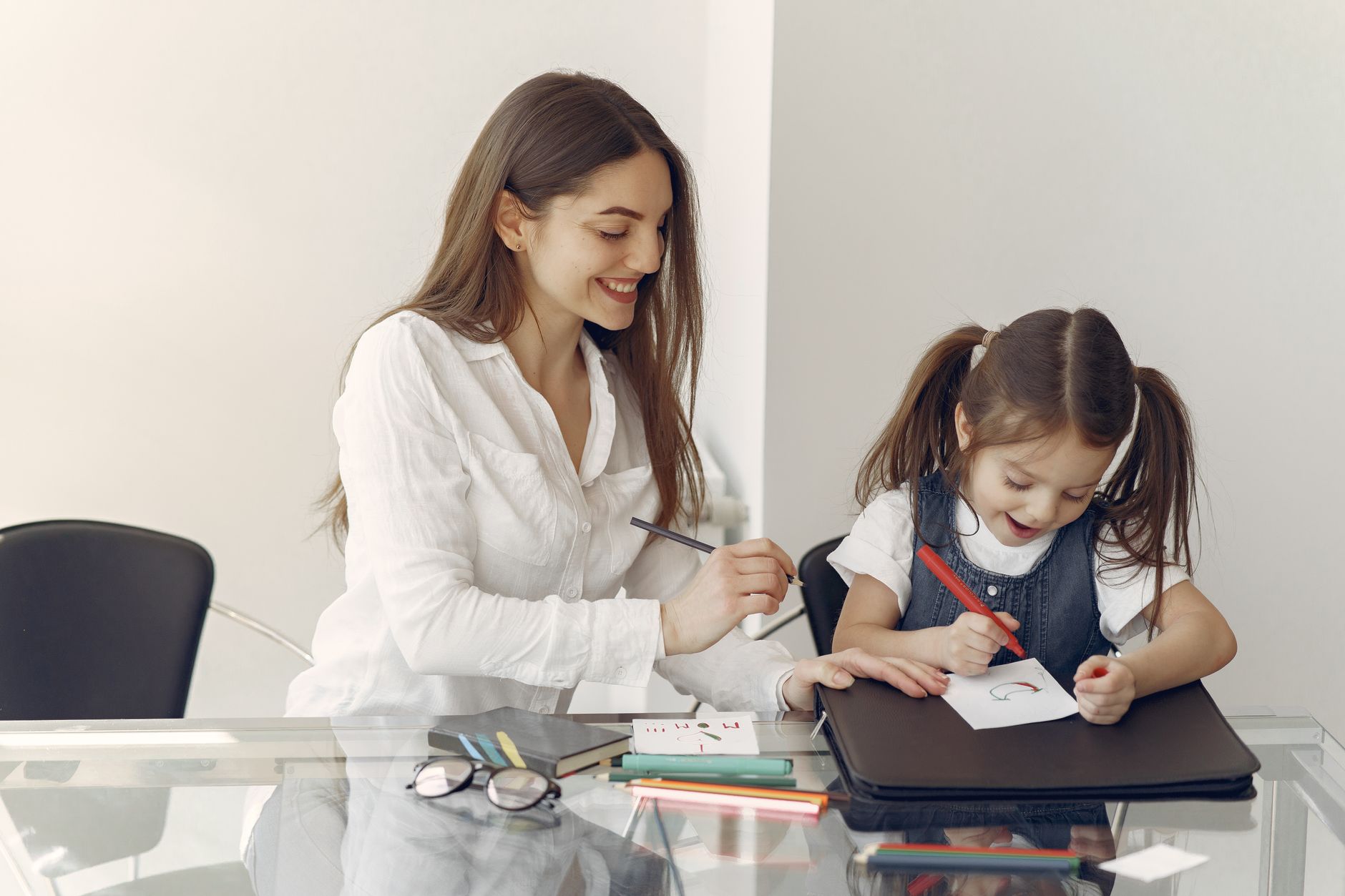 Young female tutor working with female toddler drawing together on piece of paper on document folder sitting at glass table in light office room